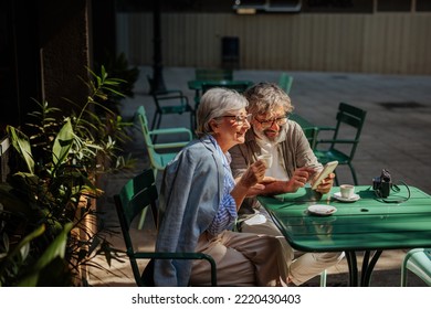 A Cheerful Senior Couple Is Outside In A Coffee Shop Garden On A Beautiful Sunny Day Having A Cup Of Coffee And Using Their Cellphone.