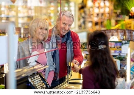 Cheerful senior couple paying with credit card at supermarket, elderly man and woman embracing, standing by cashdesk, having conversation with friendly female cashier and smiling
