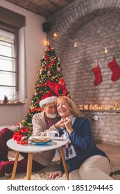 Cheerful Senior Couple Sitting By Nicely Decorated Christmas Tree, Having Fun While Drinking Coffee And Eating Gingerbread Cookies On Christmas Morning, Laughing And Hugging