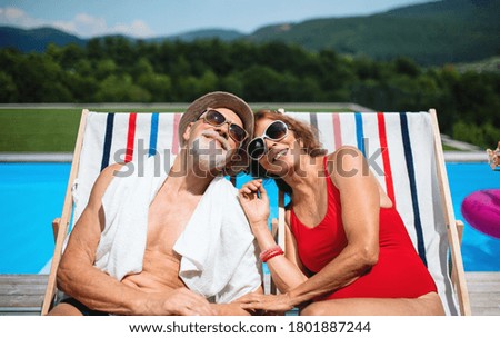 Cheerful senior couple sitting by swimming pool outdoors in backyard.