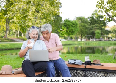 Cheerful senior couple sitting on a bench in park, Asian senior man and woman use laptop and digital tablet in park - Powered by Shutterstock