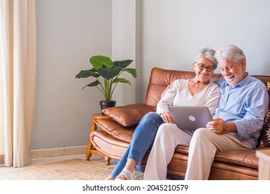 Cheerful senior couple using laptop while sitting on sofa and smiling. Elderly happy couple relaxing while surfing on laptop sitting in living room. Old couple watching media content using laptop - Powered by Shutterstock
