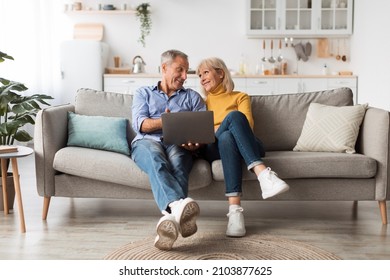 Cheerful Senior Couple Using Laptop Computer Sitting On Couch And Browsing Internet Together At Home. Mature People Use Gadget Watching Movie Online On Weekend. Full Length Shot