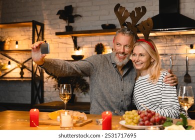 Cheerful senior couple wearing deer`s hat taking selfie during Christmas dinner in the kitchen. Merry Christmas and Happy New Year! Quality time together on celebration - Powered by Shutterstock