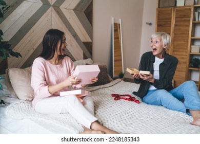 Cheerful senior diverse friends sitting on cozy bed in bedroom enjoying gifts surprisingly looking at each other spending holiday time together - Powered by Shutterstock