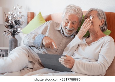 Cheerful senior family couple together in bed, two elderly laughing people relaxing at home using digital tablet technology, leisure time - Powered by Shutterstock