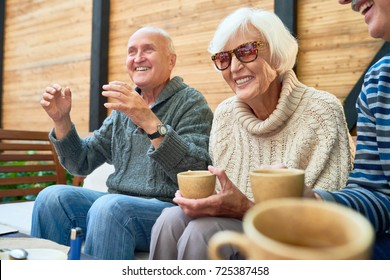 Cheerful Senior Friends Gathered Together At Cozy Small Patio And Remembering Funny Stories From Their Past, They Wearing Knitted Sweaters And Warming Themselves With Herbal Tea