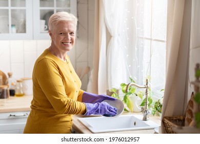 Cheerful Senior Grey-haired Lady Washing Dishes In Cozy Kitchen, Using Rubber Gloves To Protect Her Hands, Back View, Copy Space. Attractive Grandmother Dishwashing At Home, Cleaning Plate With Cloth