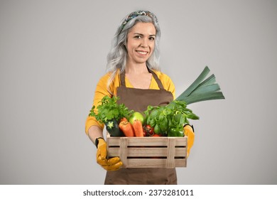 Cheerful senior lady in apron with gray hair holds box with fresh vegetables, isolated on gray background. Healthy food and harvest, work in greenhouse, farmer, agriculture - Powered by Shutterstock