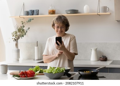 Cheerful senior lady reading online recipe book om smartphone, making video call, preparing dinner in kitchen, using mobile phone at table with healthy organic food, dish, meals, pan - Powered by Shutterstock