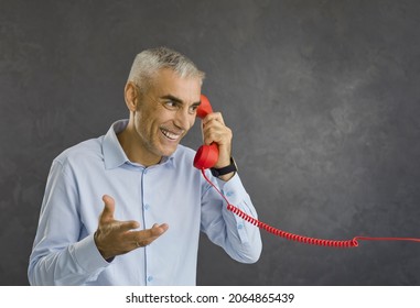 Cheerful Senior Man Answering Phone Call. Portrait Of Happy Charismatic Handsome Aged Pensioner Talking On Red Fixed Land Line Telephone, Smiling And Gesturing Standing Against Grey Studio Background