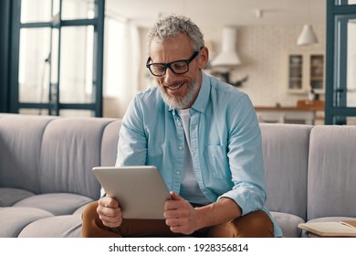 Cheerful senior man in casual clothing using digital tablet while sitting on the sofa at home - Powered by Shutterstock