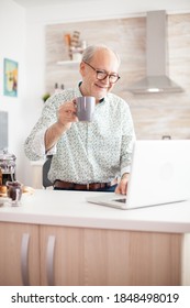 Cheerful Senior Man During Video Conference In Kitchen On The Laptop While Enjoying Breakfast And A Cup Of Coffee. Elderly Person Using Internet Online Chat Technology Video Webcam Making A Video Call