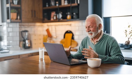 Cheerful senior man during video conference in kitchen on the laptop while enjoying breakfast and a cup of coffee. Elderly person using internet online chat technology video webcam making a video call - Powered by Shutterstock