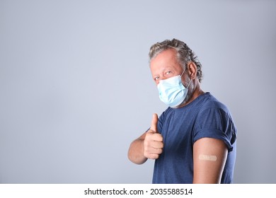 Cheerful Senior Man In Protective Mask Showing Arm With Bandage After Vaccination On Grey Background. Space For Text