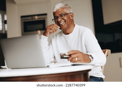 Cheerful senior man smiling at the camera while shopping online at home. Happy mature man using his credit card to place an online order on a laptop. Senior man spoiling himself after retirement. - Powered by Shutterstock