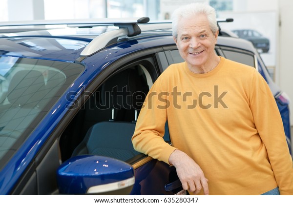 Cheerful senior man smiling happily posing near a\
new car at the dealership showroom copyspace consumerism purchasing\
buying renting leasing owning ownership customer retiring offer\
discount concept