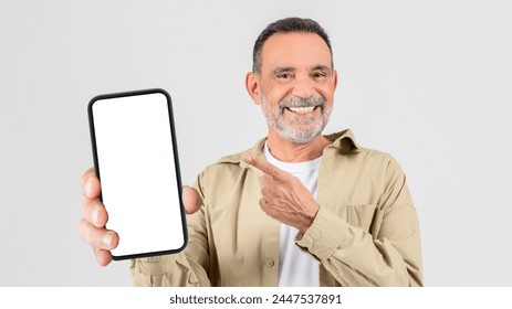 A cheerful senior man with stubble in a casual shirt holding up a smartphone with a blank white screen, pointing at it with his other hand - Powered by Shutterstock