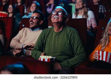 A cheerful senior man is in the theater watching a movie, smiling and eating popcorn. - Powered by Shutterstock