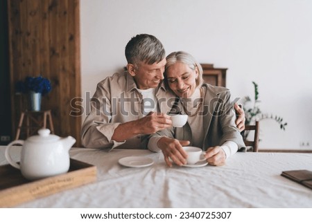 Cheerful senior man and woman in casual clothes enjoying coffee while spending romantic time together at table during breakfast at home
