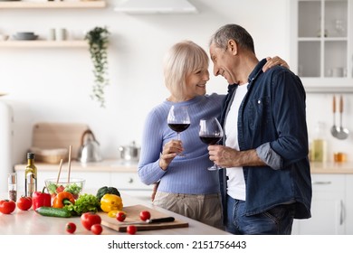 Cheerful Senior Man And Woman Hugging And Clinking With Red Wine Glasses In Kitchen Interior, Happy Beautiful Elderly Husband And Wife Cooking Dinner Togeher, Side View, Copy Space