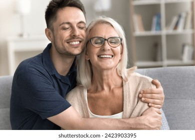 Cheerful senior mom and son man enjoying leisure, family closeness, meeting at home, sitting on couch, hugging with heads touch, laughing with closed eyes. Adult male child embracing mother with care