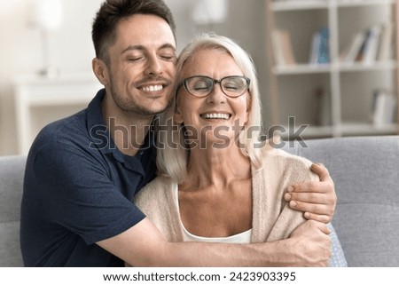 Cheerful senior mom and son man enjoying leisure, family closeness, meeting at home, sitting on couch, hugging with heads touch, laughing with closed eyes. Adult male child embracing mother with care