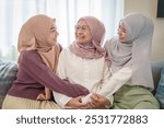 cheerful senior mother and adult daughters in hijab dresses sitting on sofa,embracing together,smiling and looking each other,family lifestyle,meeting and relationship