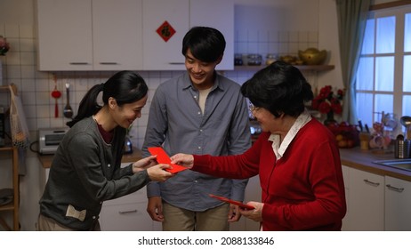 Cheerful Senior Mother Giving Red Envelopes Lucky Money To Surprised Adult Son, Daughter In Law And Granddaughter With Hand Gestures After Dinner On Chinese New Year's Eve