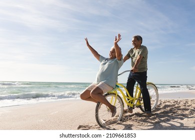 Cheerful Senior Multiracial Couple Enjoying Retirement With Bicycle At Beach On Sunny Day. Active Lifestyle And Transportation.