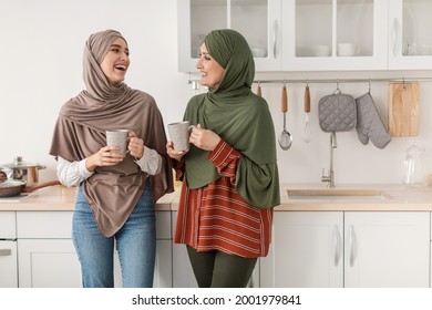 Cheerful Senior Muslim Mother And Adult Daughter Wearing Hijab Enjoying Morning Coffee Talking And Laughing Together Standing In Modern Kitchen At Home. Happy Middle-Eastern Family Concept
