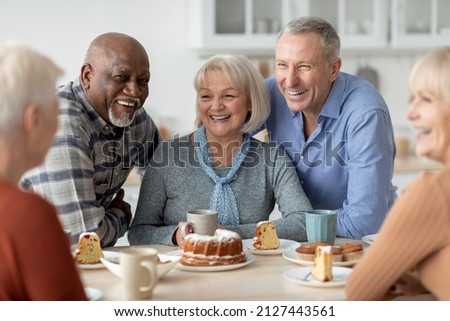 Cheerful senior people of different nationalities sitting at kitchen, drinking tea and eating cake together, having conversation and laughing, chilling together at nursing home
