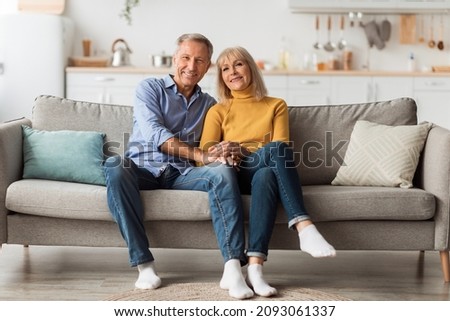 Cheerful Senior Spouses Holding Hands Hugging Sitting On Sofa Smiling To Camera At Home. Front View Of Happy Married Mature Couple Posing On Sofa. Long-Lasting Marriage And Relationship