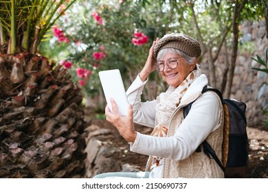 Cheerful Senior Woman With Cap And Backpack In Outdoors Excursion Sitting Video Chatting With Digital Tablet.  Elderly Attractive Female Resting In Shadow Of Tropical Woods