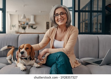 Cheerful senior woman in casual clothing spending time with her dog while sitting on the sofa at home