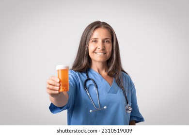 Cheerful senior woman doctor in uniform showing jar of pills to camera, posing on grey studio background. Professional recommendation, treatment, health care and medicine - Shutterstock ID 2310519009