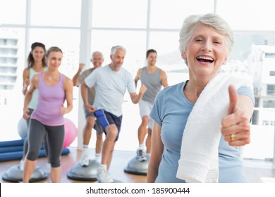 Cheerful senior woman gesturing thumbs up with people exercising in the background at fitness studio