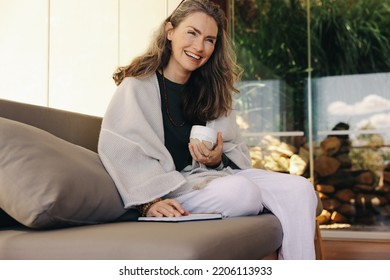 Cheerful Senior Woman Journaling With A Cup Of Tea In The Morning. Mature Woman Smiling Happily While Sitting On A Couch. Senior Woman Enjoying Relaxing In Her Living Room At Home.