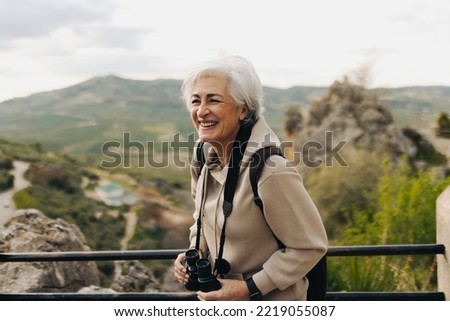 Cheerful senior woman laughing while standing on a hilltop with binoculars. Happy elderly woman enjoying a leisurely hike outdoors. Mature woman enjoying recreational activities after retirement.