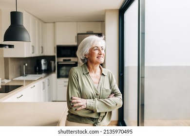 Cheerful senior woman looking away thoughtfully while standing in her home. Happy elderly woman smiling while reflecting on memories of the past. Stock-foto