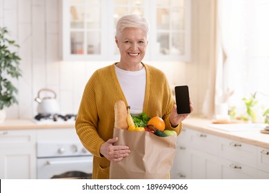 Cheerful Senior Woman With Shopping Bag Full Of Grocery Showing Smartphone With Blank Screen, Kitchen Interior, Copy Space, Mockup. Smiling Old Lady Using Mobile App For Grocery Delivery