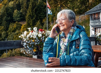 Cheerful Senior Woman Sitting At Restaurant Table Outdoors In The Mountain Talking On Mobile Phone While Holding A Coffee Cup. Elderly Traveler Lady Enjoying Freedom And Retirement