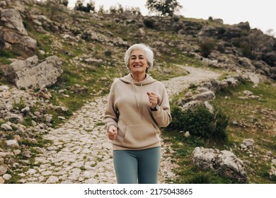 Cheerful Senior Woman Smiling While Jogging Alone Outdoors. Happy Elderly Woman Working Out On A Hilly Trail. Mature Woman Maintaining A Healthy Lifestyle After Retirement.