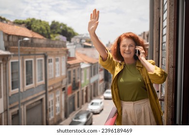 Cheerful senior woman standing on balcony, waving hand and talking on cellphone, happy friendly elderly lady raising arm, greeting somebody and enjoying mobile phone conversation, copy space - Powered by Shutterstock