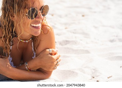Cheerful sexy young woman in sunglasses, beads necklace and rings lying down on sand at beach and looking away during summer holiday. Woman relaxing on sand at beach on a bright sunny day