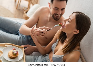 Cheerful Shirtless Man Feeding Sexy Girlfriend With Fresh Strawberry During Breakfast In Bedroom