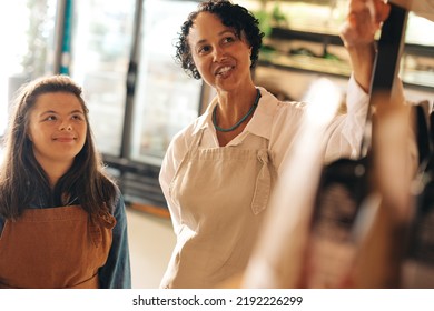 Cheerful Shop Supervisor Giving Instructions To Her New Employee In A Grocery Store. Female Small Business Owner Employing A Woman With Down Syndrome As A Retail Sales Worker.