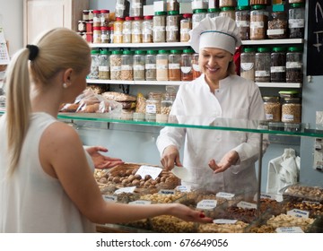 Cheerful Shop Woman Offering Senior Client Nuts In Local Confectionery