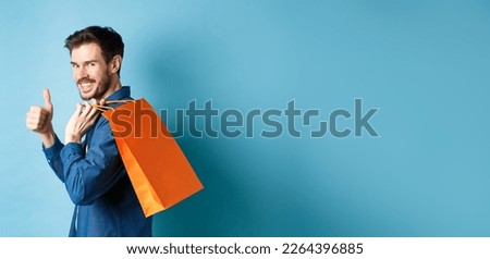 Cheerful shopper holding orange shopping bag on shoulder, turn around at camera with thumbs up, recommending store, blue background.