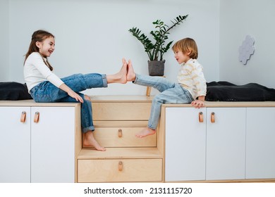 Cheerful siblings sitting on wooden elevation in front of each other , presing bare feet for fun. Side view. - Shutterstock ID 2131115720
